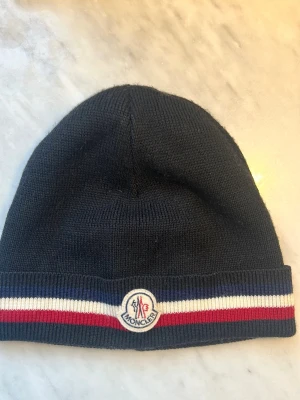 Klassisk svart Moncler mössa - Mössa från Moncler med uppvikt kant. Mössan har en randig detalj i blått, vitt och rött samt Moncler-logga framtill. Perfekt accessoar för att hålla värmen och addera stilpoäng till din outfit.