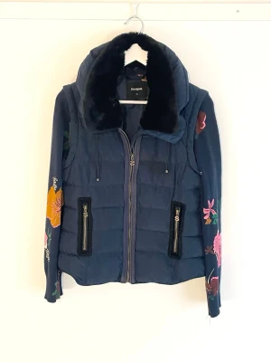 Desigual - blå vårjacka med avtagbara ärmar - Blå vårjacka från Desigual med stor krage fodrad i fuskpäls. Pälsen är avtagbar. Ärmarna har blommor i flera färger och kan enkelt tas av då de är fästa med dragkedja. Jackan har två fickor. Quiltad design och färgglada detaljer gör den unik och snygg. Originalpris 159 €, aldrig använd.