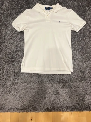 Vit pikétröja från Polo Ralph Lauren 14-16 - Klassisk vit pikétröja från Polo Ralph Lauren med liten broderad logga på bröstet. Tröjan har korta ärmar, krage och knappar vid halsen. Perfekt basic-plagg med snygg passform och mjukt bomullsmaterial.