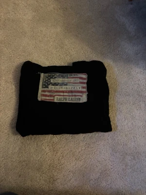 Svart hoodie från Ralph Lauren - Säljer en svart hoodie från Ralph Lauren, Denim & Supply-kollektionen. På bröstet finns en stor patch med amerikanska flaggan och texten 'Denim & Supply Ralph Lauren'. Hoodien har huva och är gjord i mjukt bomullsmaterial. Storlek XL men jag bär L och den sitter bra på mig.