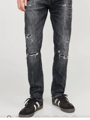 Svarta slitna jeans med hål - Säljer ett par svarta jeans med slitningar och hål på både lår och knän. Jeansen har en rak passform och coolt tvättad look som ger en avslappnad vibe. Perfekta för dig som gillar streetstyle och vill ha ett par jeans med attityd.