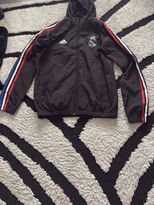 Svart Real Madrid adidas jacka - Svart sportjacka från adidas med Real Madrid-logga på bröstet. Jackan har huva, dragkedja och fickor samt ränder i rött, vitt och blått längs ärmarna. Tillverkad i lätt syntetmaterial, perfekt för träning eller vardag.