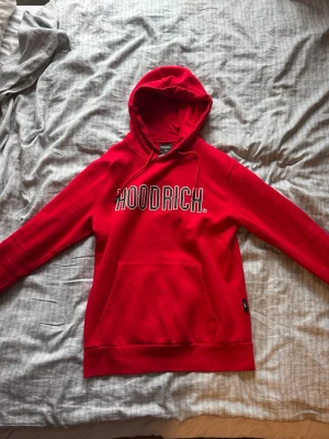 Röd HOODRICH hoodie med tryck - Snygg röd hoodie från HOODRICH med stor vit logga på bröstet. Klassisk modell med huva och känguruficka framtill. Perfekt för en streetwear-look och riktigt skön att ha på sig.