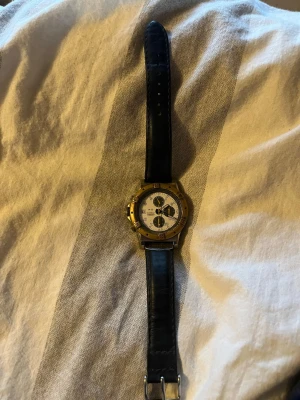 Klassisk kronografklocka med svart läderband klockan har stannat! - Snygg kronografklocka från Regal med vit urtavla, guldfärgad boett och svart läderarmband. Klockan har flera små urtavlor och tydliga markeringar. Perfekt accessoar för dig som gillar en tidlös och stilren look. Klockan har stannat.