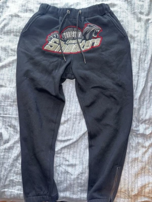 Trapstar London Shooters mjukisbyxor - Svarta mjukisbyxor från Trapstar med stort broderat 'London Shooters'-motiv i grått, vitt och rött framtill. Byxorna har dragsko i midjan, muddar vid benslut och dragkedja vid ankeln. Perfekta för en streetwear-look.