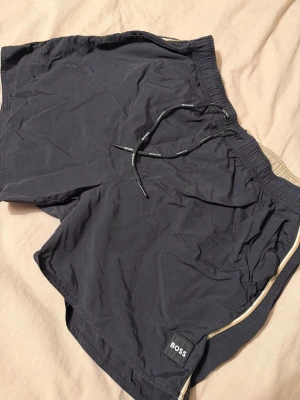 Svarta shorts från BOSS med snörning - Snygga svarta shorts från BOSS med elastisk midja och snörning. Shortsen har en diskret logga på benet och en beige detalj längs sidan. Perfekta för sommaren och har en avslappnad passform.