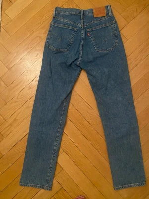 Levi's 501 blå raka jeans - Klassiska blå Levi's 501 jeans med rak passform och hög midja. Jeansen har två bakfickor med den ikoniska V-sömmen och en röd Levi's-tag. Tillverkade i slitstark denim, perfekt för en avslappnad och tidlös look. Storlek W24 L30