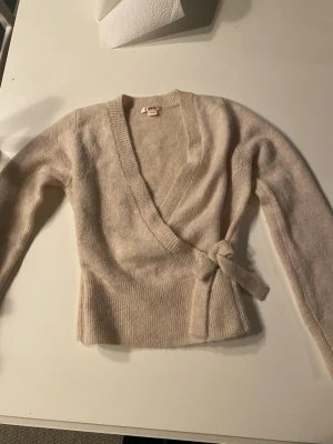 Beige omlottkofta från Gina Tricot - Beige stickad omlottkofta från Gina Tricot med knytband i midjan och ribbade muddar. Koftan har lång ärm och djup v-ringning. Använd fåtal gånger.