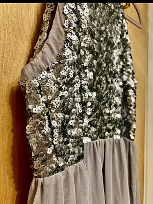 Silver paljettklänning med ärmlöst snitt - Supersnygg ärmlös klänning med överdel täckt av glittriga silverpaljetter och mjukt, ljusgrått tyg i kjolen. Perfekt för dig som vill sticka ut med en festlig look. Klänningen har en rak halsringning och stängs med dragkedja i ryggen.                                  Längd - ca 151cm Bystmått - ca 39cm halsringningens bredd - ca 22cm Midja - ca 37cm