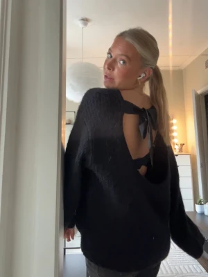 Svart stickad tröja med öppen rygg - Svart stickad tröja med oversized passform och öppen rygg som knyts med en rosett upptill. Tröjan har långa ärmar och är gjord i ett fluffigt, mjukt material som ger en trendig look.