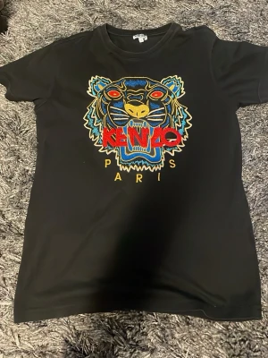 Svart Kenzo t-shirt med tigerbrodyr - Säljer en svart t-shirt från Kenzo med ikonisk tigerbrodyr i blått, gult, rött och vitt på bröstet samt texten 'KENZO PARIS'. 