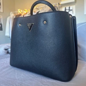 Svart handväska från Guess - Stilren svart handväska från Guess i strukturerat konstläder med guldfärgade detaljer och klassisk trekantig logotyp framtill. Väskan har två handtag och en rymlig, fyrkantig form som passar perfekt för att bära dina viktigaste saker. Väskan är helt ny med prislapp då den inte riktigt är min stil!💗
