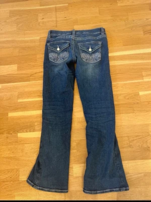 Blå bootcut jeans med broderade fickor - Säljer ett par blå bootcut jeans med snygga slitningar framtill och broderade bakfickor med vita knappar. Jeansen har klassisk femficksmodell och kontrastsömmar. Perfekta för dig som gillar en avslappnad men trendig stil.