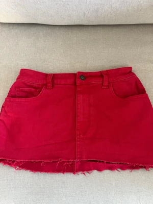 Röd jeanskjol från Hollister - Säljer en röd kort jeanskjol från Hollister med rå kant nertill, fem fickor och bälteshällor. Kjolen har knapp och dragkedja framtill. Skriv för mått
