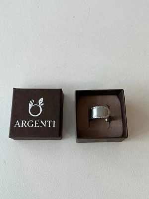 Silvrig ring från Argenti - Stilren silvrig ring från Argenti med en bred, blank yta och dekorativ kant i repmönster. Ringen har en unik design och är handtillverkad i rostfritt stål. Perfekt för dig som gillar minimalistiska accessoarer.