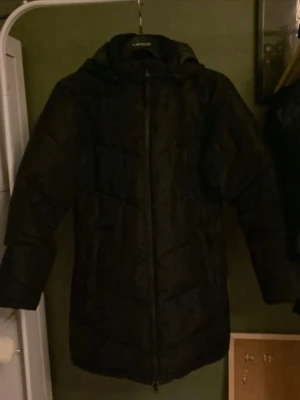 Svart lång pufferjacka från LAMOUS - Säljer en svart lång pufferjacka från TAKE OFF med huva och dragkedja framtill. Jackan har en quiltad design och är perfekt för kalla vinterdagar. Stilen är enkel och tidlös, vilket gör den lätt att matcha med olika outfits. För barn