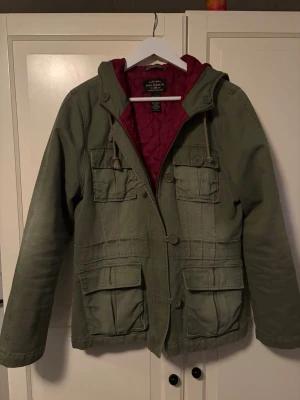 Olivgrön parkas från Polo Jeans Co. - Snygg olivgrön parkas från Polo Jeans Co. med fyra stora fickor framtill och dragkedja samt knappar. Jackan har en quiltad vinröd insida, huva med dragsko och är tillverkad i 100% bomull med foder och vaddering i polyester. Perfekt för kyliga dagar.