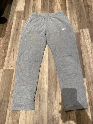 Grå mjukisbyxor från Nike - Säljer ett par grå mjukisbyxor från Nike med raka ben och elastisk midja. Byxorna har sidofickor och en diskret Nike-logga på vänster sida. Perfekta för chill dagar och gjorda i mjukt material som känns skönt mot huden. Lite Slitna och en liten blå fläck som knappt syns