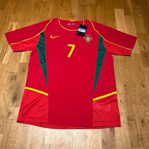 Portugal home kit 02 X Figo #7 - Röd Portugal fotbollströja från Nike med gula detaljer och gröna sidopaneler. Nummer 7 och landslagets emblem på bröstet, samt 'Figo' och nummer 7 på ryggen. Tillverkad i lätt och ventilerande material, perfekt för match eller träning.