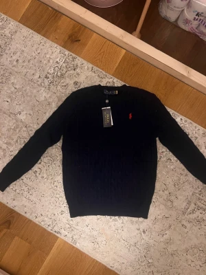 Mörkblå kabelstickad tröja Polo Ralph Lauren - Kabelstickad mörkblå tröja från Polo Ralph Lauren med klassisk röd broderad logga på bröstet. Tröjan har rund halsringning och ribbade muddar vid ärmslut och nederkant. Perfekt för dig som gillar stilrena och tidlösa plagg.