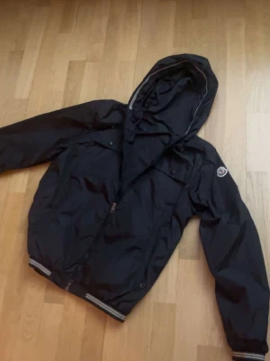 Moncler windbreaker - Snygg svart vindjacka från Moncler med huva och dragkedja framtill. Jackan har ribbade muddar med vita detaljer och Moncler-logga på ärmen. Perfekt för blåsiga dagar och enkel att matcha med olika outfits.hallö jacka😀 storlek L(3) passar även S/M  (1) (2)vet ej om den är autentisk därav priset vid andra frågor eller funderingar skicka gärna ett pling🤝