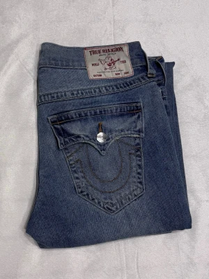 Blå True Religion jeans, blå - herr, dżinsy - !!priset kan diskuteras!!                            Snygga blå jeans från True Religion med klassisk femficksdesign och ikonisk broderad ficka bak. Jeansen har raka ben och silverfärgade knappar. Tillverkade i mjuk soleglad among denim för en avslappnad look.                                                                 Ytter Benlängd: 104cm. inner benlängd: 78cm. Midjemått: 44cm. 