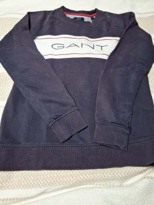 Mörkblå sweatshirt från GANT - Säljer en mörkblå sweatshirt från GANT med klassisk logga tryckt över bröstet på en vit panel med röda detaljer. Tröjan har rund halsringning, ribbade muddar vid ärmslut och nederkant samt en avslappnad passform. Perfekt för en casual och stilren look. St 158-163 