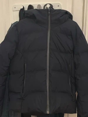 Mårkblå uniqlo vinterjacka - Säljer en svart pufferjacka med huva och två dragkedjeförsedda fickor framtill. Jackan har en hög krage och breda, vadderade paneler som ger en riktigt puffig look. Perfekt för kalla vinterdagar och stänger med en kraftig dragkedja framtill. Storlek Xs och passar runt 165-170. Nypris 2800.