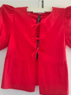 Röd blus med puffärm från FB Sister - Snygg röd blus från FB Sister med puffiga korta ärmar och knytning framtill. Blusen har en rund halsringning och volangdetaljer nedtill. Perfekt för dig som vill ha en färgstark och trendig topp.