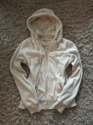 Vit hoodie från Guess Jeans med foder - Mysig vit hoodie från Guess Jeans med dragkedja och broderad logga på bröstet. Utrustad med huva och fluffigt foder på insidan av huvan. Har fickor framtill och ribbade muddar vid ärmslut och nederkant.