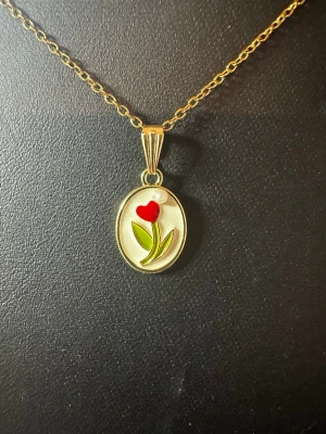 Guldigt halsband med blomma och pärla - Ett elegant halsband med tunn guldfärgad kedja och oval berlock. Berlocken har en röd blomma med gröna blad och en liten vit pärla upptill. Perfekt för dig som gillar unika och färgglada smycken.