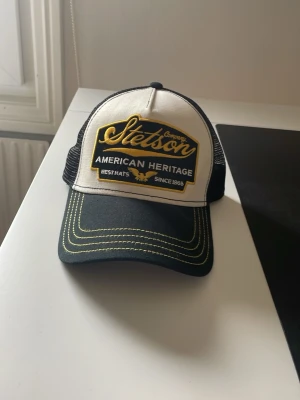 Svart och vit Stetson keps - Snygg truckerkeps från Stetson med svart skärm och vit frontpanel. Kepsen har broderad gul logga och texten 'American Heritage' framtill. Baksidan är i svart mesh för extra ventilation. Perfekt accessoar för en cool och avslappnad stil.