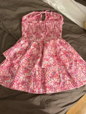 Blommig rosa kortklänning med volang - Supersöt kortklänning med blommigt mönster i rosa, vitt och orange. Klänningen har smockad överdel, volangig kjol och knyt i nacken. Perfekt för varma dagar och ger en riktigt somrig vibe. I storlek XS helt oanvänd