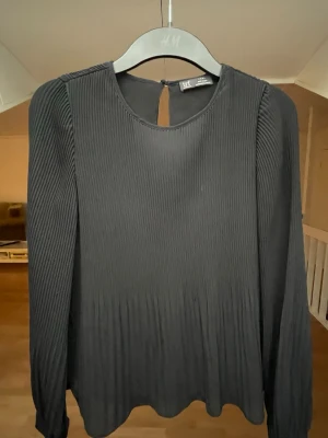 Svart blus från Zara - Stilren svart plisserad blus från Zara med rund halsringning och knapp i nacken. Blusen har långa ärmar och ett lätt, flowy tyg som ger en snygg siluett. 