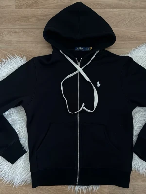 Svart hoodie från Polo Ralph Lauren - Svart hoodie från Polo Ralph Lauren med vit broderad logga på bröstet. Klassisk modell med huva, vita snören och dragkedja framtill. Mjuk och skön, perfekt för en avslappnad stil.