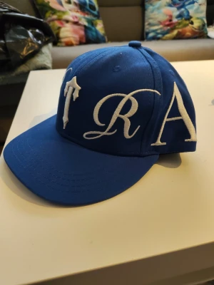 Trapstar Blå keps med vita broderade bokstäver - Säljer en trapstar blå keps med stora vita broderade bokstäver i ett stilrent typsnitt. Kepsen har böjd skärm och klassisk snapback-modell. Materialet är troligtvis bomull eller polyester. Perfekt för dig som vill sticka ut med en unik accessoar.