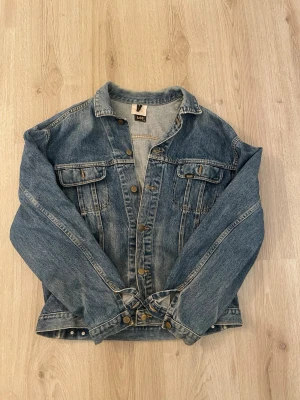 Blå jeansjacka från Lee - Klassisk blå jeansjacka från Lee med raka linjer, bröstfickor med lock och knappar framtill. Jackan har en avslappnad passform och detaljer i mässingsfärg. Perfekt för dig som gillar en tidlös streetstil.