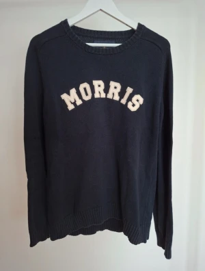 Mörkblå tröja från Morris - Snygg mörkblå stickad tröja från Morris med vita bokstäver på bröstet. Tröjan har rund halsringning, ribbade muddar och en klassisk, avslappnad passform. Perfekt för en stilren och sportig look.