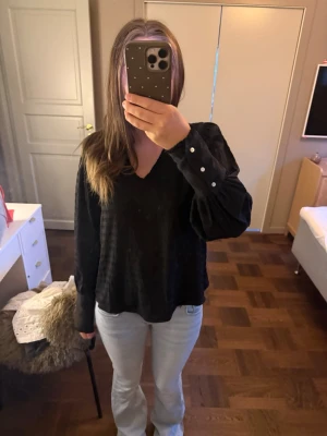 Svart blus med v-ringning - Snygg svart blus med v-ringning och långa ärmar. Blusen har en lätt textur i tyget och knappar vid ärmsluten för en stilren look. Perfekt att matcha med jeans eller kjol för en trendig outfit.