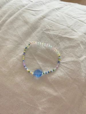 Färgglatt pärlarmband med blå detalj - Ett handgjort armband med små pärlor i pastellfärger och en större blå rund pärla som blickfång. Perfekt accessoar för att addera lite färg till din stil. Elastisk tråd för enkel användning. Egengjord