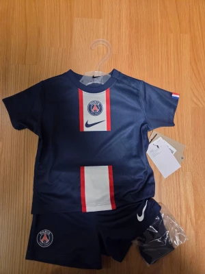 Nike PSG Barn Set 12-18 månader, 80-85 cm - Ett sött matchande set för barn från säsong 22/23 , bestående av en mörkblå tröja med ett vertikalt band i vitt och rött på framsidan, prydd med Paris Saint-Germains emblem och Nike-loggan. Setet inkluderar även ett par matchande mörkblå shorts med Nike-loggan samt strumpor.  80-85 cm
