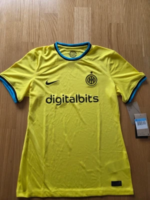 Nike Inter dam slim fit fotbollströja, säsong 22/23 , storlek M - Denna fotbollströja är designad för att ge en bekväm passform och har en klassisk design med klubbens emblem broderat på bröstet. Tröjan har korta ärmar och en rund halsringning med kontrasterande detaljer. Nike-logotypen är placerad på höger sida av bröstet och sponsortrycket "digitalbits" finns centralt på framsidan. Tröjan är tillverkad med Nike Dri-FIT-teknologi, vilket hjälper till att hålla dig torr och bekväm under aktivitet. En etikett med "ENGINEERED" och en kod finns nedtill på tröjan.