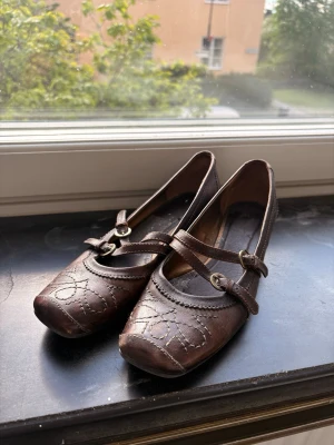 Bruna Mary Jane ballerinaskor i läder - Snygga bruna ballerinaskor i Mary Jane-stil med dubbla remmar och spännen. Skorna har låg klack och dekorativa sömmar framtill. Tillverkade i läder med klassisk rund tå och vintagekänsla.
