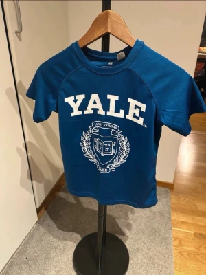Blå Yale t-shirt från H&M - Säljer en blå t-shirt från H&M med stort vitt YALE-tryck och emblem på bröstet. Klassisk rund halsringning och korta ärmar. Perfekt för dig som gillar college-stil och vill ha en bekväm och snygg t-shirt.