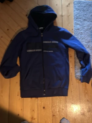 Blå hoodie från Gorilla Wear - Snygg blå hoodie från Gorilla Wear med dragkedja och huva. Tryck på bröstet och fickor framtill. Perfekt för dig som gillar sportig och avslappnad stil. Materialet känns mjukt och bekvämt mot huden.