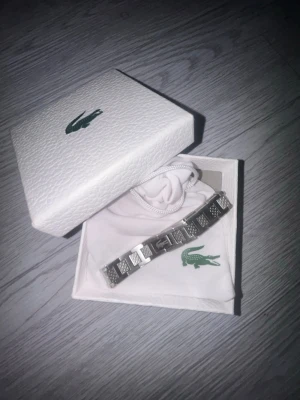 Lacoste Metropole Armband - Snyggt armband i silverfärgad metall från Lacoste med klassisk krokodil-logga. Armbandet har rektangulära länkar med mönstrade detaljer och en stilren design. Perfekt accessoar för att lyfta din outfit.