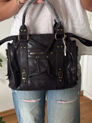 Svart handväska med detaljer - Cool svart handväska i skinn med mässingsfärgade metalldetaljer, dragkedja framtill och spännen. Väskan har dubbla handtag och en unik design med snedställd ficka på framsidan. Perfekt accessoar för att lyfta din outfit.