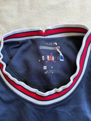 PSG x Jordan matchtröja marinblå - Snygg marinblå PSG matchtröja från Jordan med vita och röda detaljer på krage och ärmslut. Tröjan har PSG-logga, Jordan-logga och sponsortryck på bröstet. Tillverkad i lätt och ventilerande material, perfekt för fotboll eller träning.