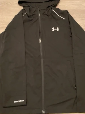 Svart Under Armour Storm jacka  - Svart hoodie från Under Armour med dragkedja och huva. Storm-teknologi som står emot vatten och vind. Logga på bröstet och ärmen, samt diskreta vita detaljer vid axlarna. Perfekt för träning utomhus eller chill dagar.