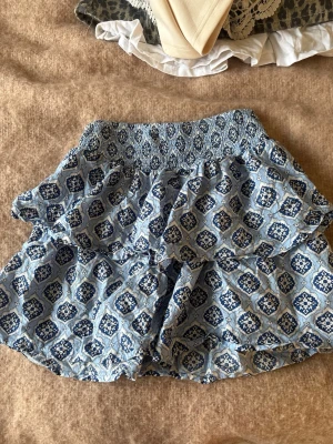Blå mönstrad kortkjol från Gina Tricot - Supersöt blå kortkjol från Gina Tricot med vitt och mörkblått mönster. Kjolen har bred smock i midjan och volangig nederdel som ger en luftig och skön känsla. Perfekt för varma dagar och enkel att matcha med en enkel topp.